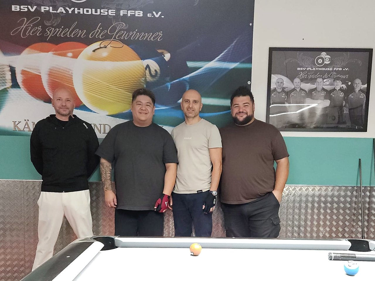 Vier lächelnde Männer stehen im Vereinsheim des BSV Playhouse FFB hinter einem Pool-Billardtisch, an der Wand ein Banner mit dem Vereinsnamen und gerahmte Mannschaftsfotos